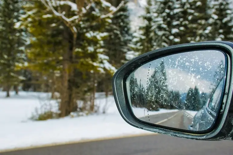 Snow-covered car side mirror reflecting a winter road lined with trees