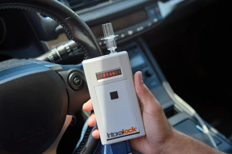 A person holding an Intoxalock ignition interlock device in front of a car's steering wheel, with the device's display showing the message 'Blow'