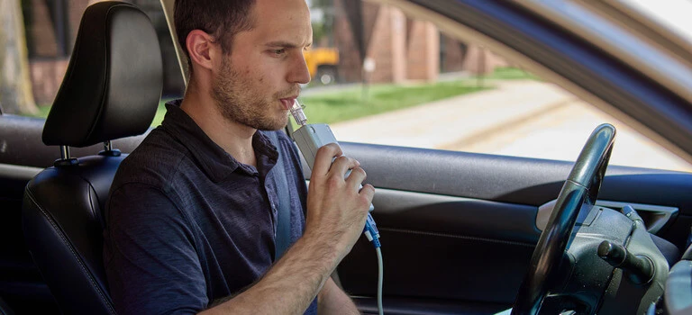 Man sitting in a car using an ignition interlock device, blowing into an Intoxalock ignition interlock device to provide a breath sample.