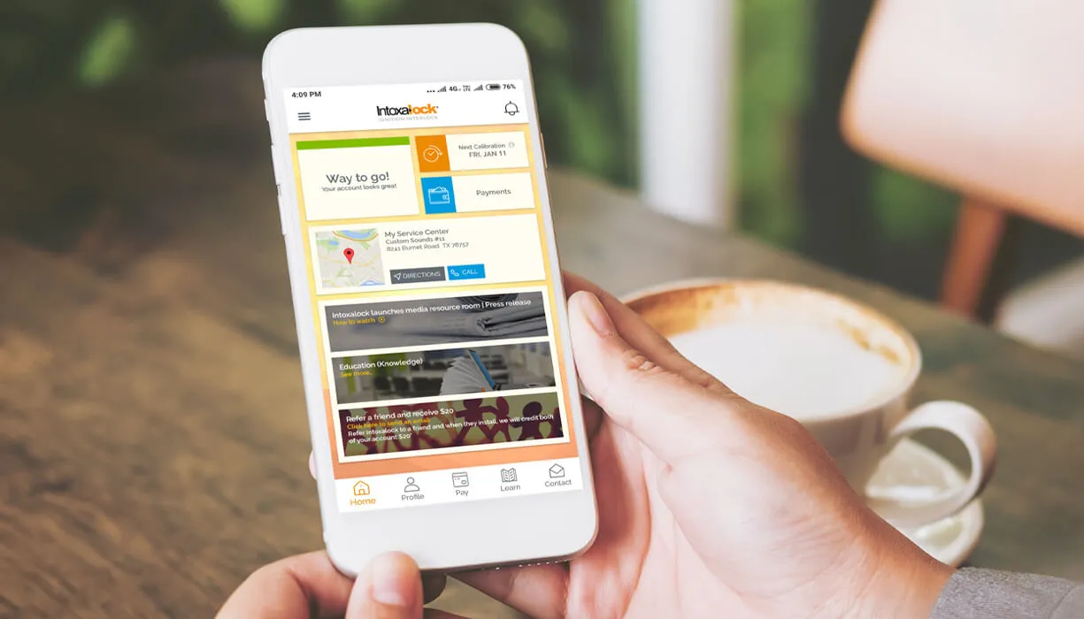 Hand holding a smartphone with the Intoxalock app, showing account management options like payments, service center, and resources for ignition interlock users