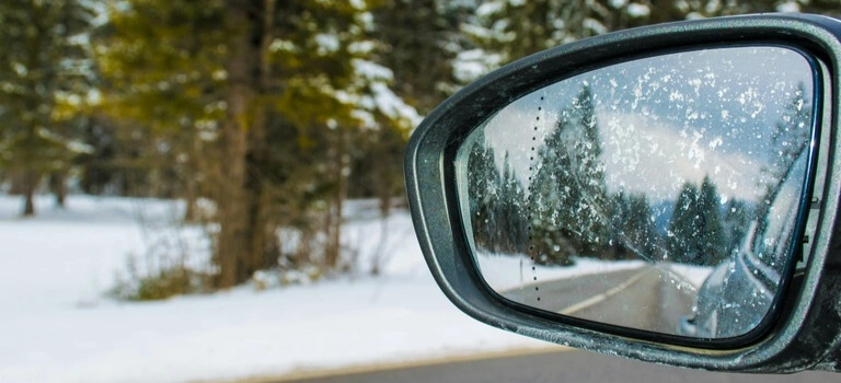 Snow-covered car side mirror reflecting a winter road lined with trees