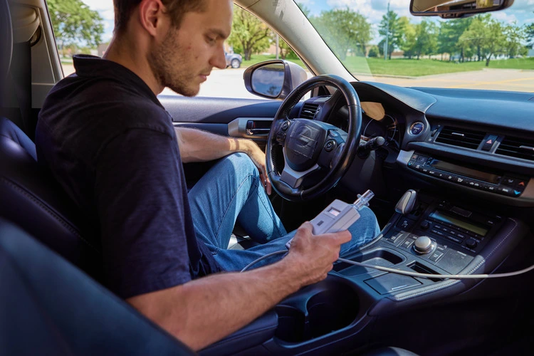 A man using an Intoxalock ignition interlock device in a car, preparing to take a breath sample.