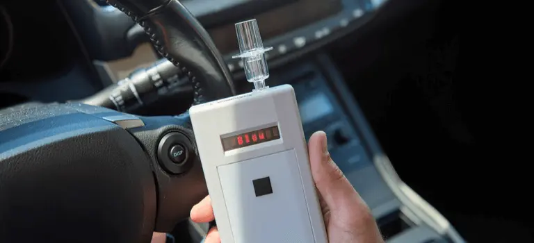 A person holding an Intoxalock ignition interlock device in front of a car's steering wheel, with the device's display showing the message 'Blow,'