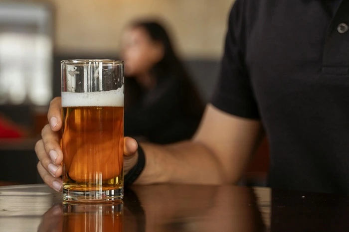 Person holding a glass of beer on a bar counter, with another patron blurred in the background