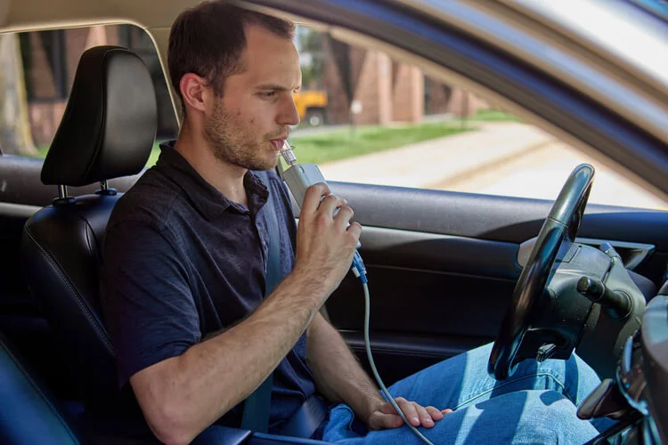 Man sitting in a car using an ignition interlock device, blowing into an Intoxalock ignition interlock device to provide a breath sample.